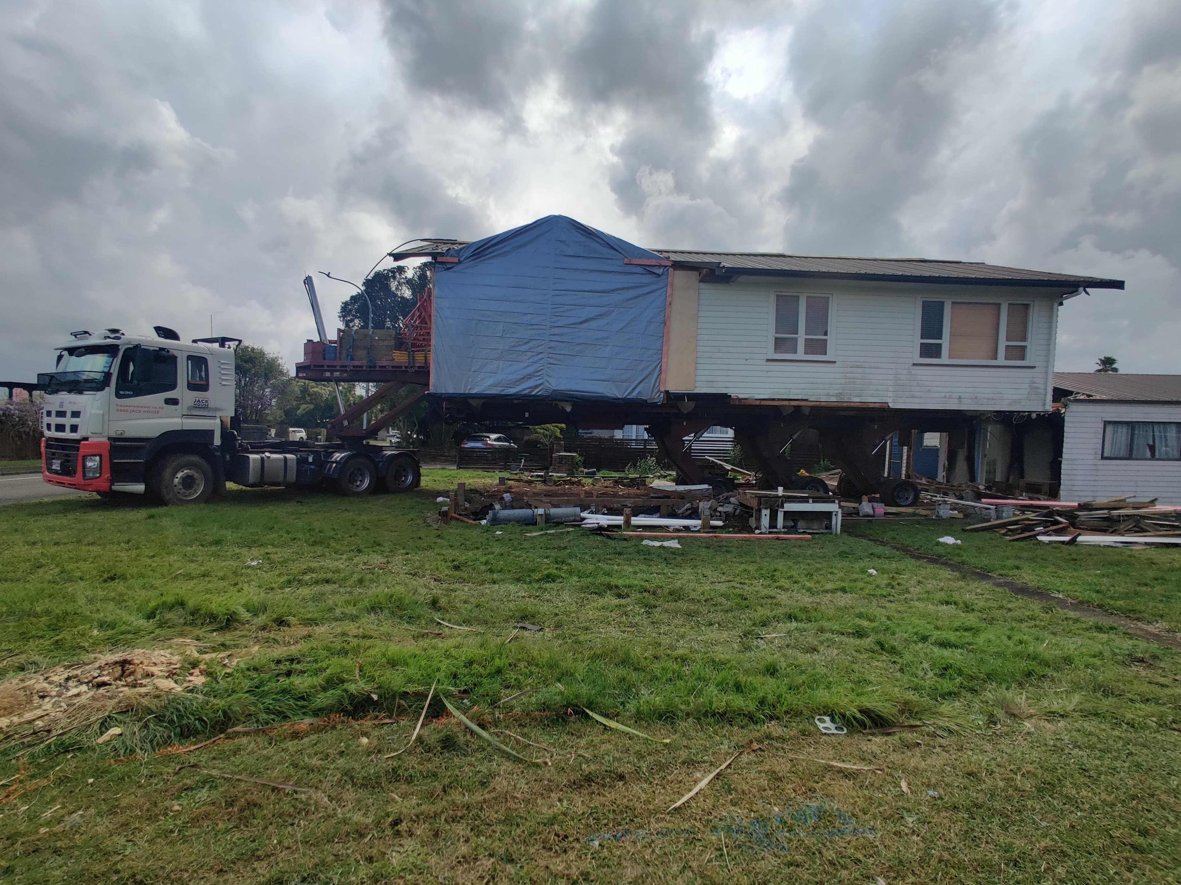 A weatherboard house section on a heavy haulage trailer behind a semi-truck — the overnight road move stage of a relocatable home project.
