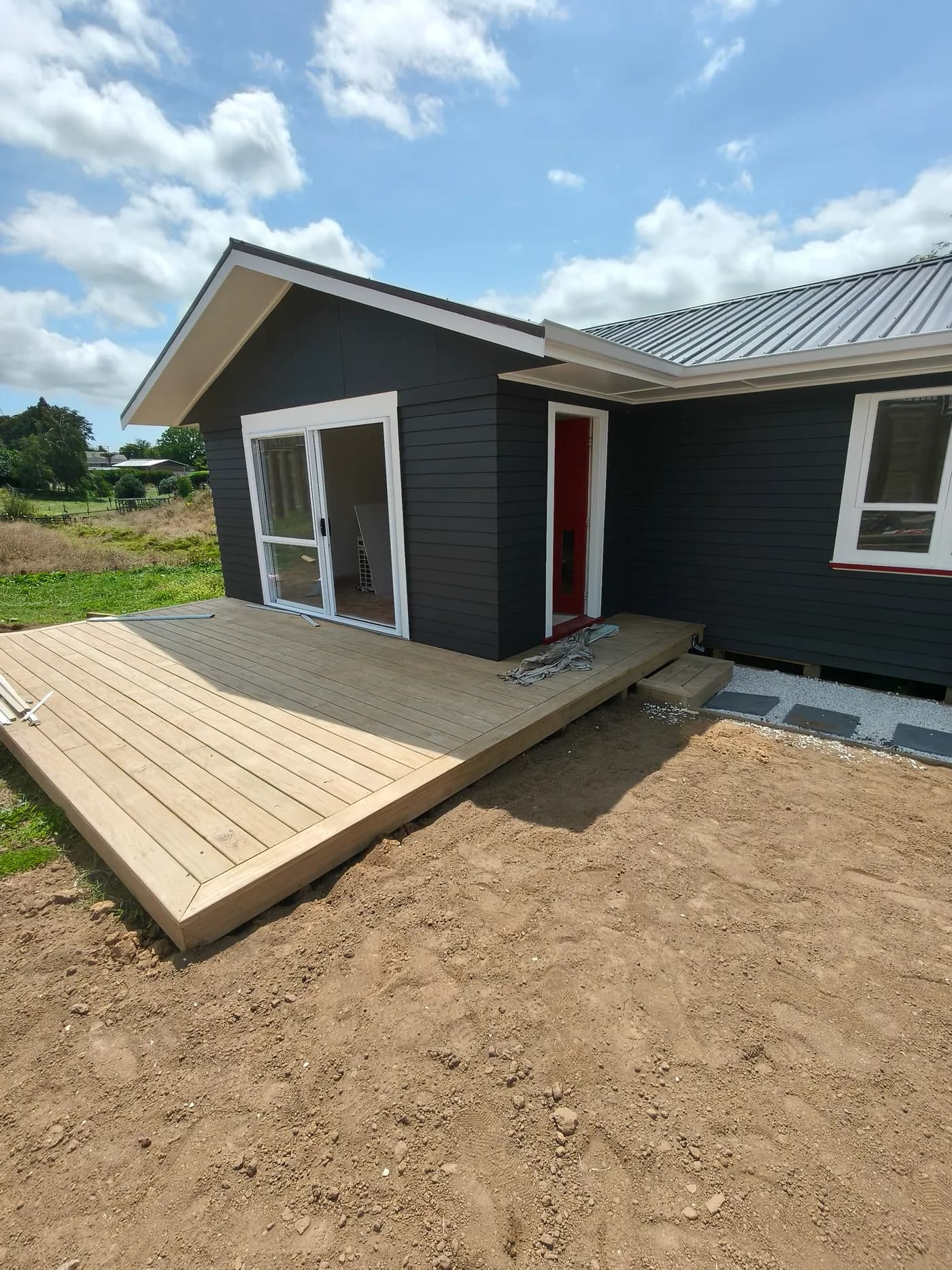 Modern relocatable-style home with charcoal cladding, timber deck, and red front door on a rural section.