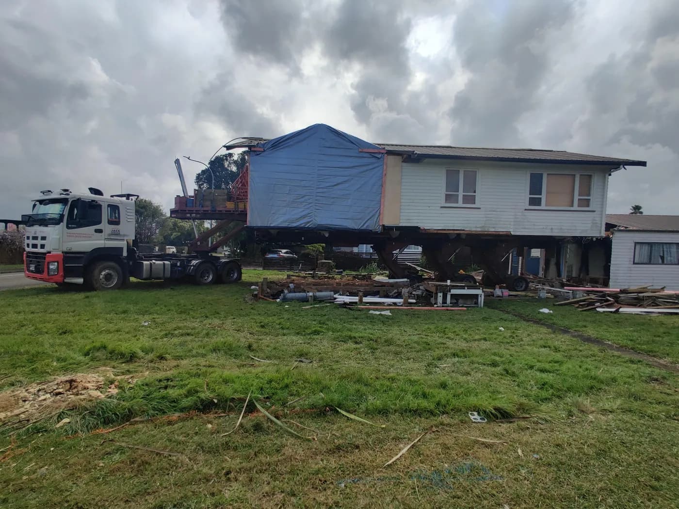 A weatherboard house section on a heavy haulage trailer behind a semi-truck — the overnight road move stage of a relocatable home project.